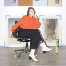 a white woman in her 30s seated on a black rolling chair. shes's wearing an orange sweater, black pants, and white boots. art works are on the wall behind her.
