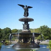 A large bronze fountain featuring an angelic figure with wings outstretched atop a tiered pedestal, water cascading gently into a circular pool below. The statue is surrounded by lush green trees under a bright blue sky, with aquatic plants in the foreground and people visible in the distance along the water’s edge.