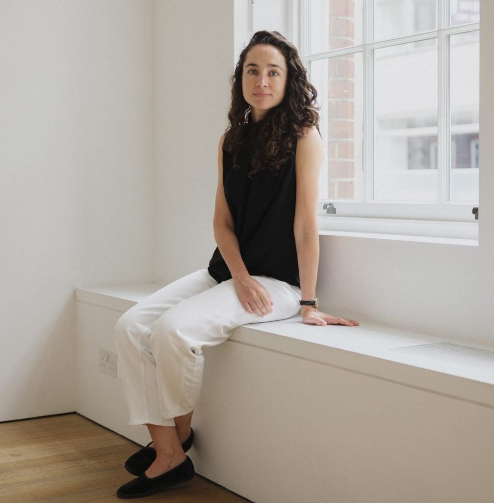 A woman in a black top and white pants sits on a ledge against a window
