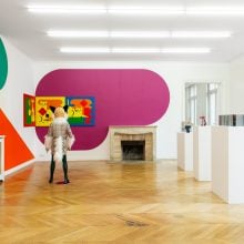 Contemporary art exhibition featuring colorful geometric wall paintings, abstract sculptures on white pedestals, and a visitor wearing a fox costume in a bright gallery space.