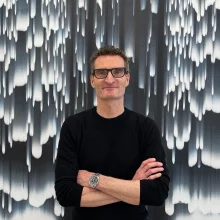 Stanley Casselman with short hair and glasses stands confidently with his arms crossed in front of an abstract black-and-white artwork featuring vertical, dripping light patterns. He is wearing a black sweater and a wristwatch, and he is smiling slightly, looking directly at the camera.