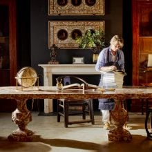 A woman polishing furniture pieces on a large marble table in an old house that has a white fire place and lots of ornaments.