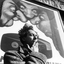 A black-and-white photograph of a man smoking a cigarette in front of a large illustrated poster depicting exaggerated faces with long noses peering downward. The man stands in shadow, his eyes closed, framed against the bold and surreal background imagery. Featured in a surrealism exhibition at Throckmorton Fine Art.