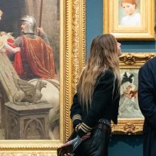 A young woman and man look at paintings in gold frames hung on a blue wall