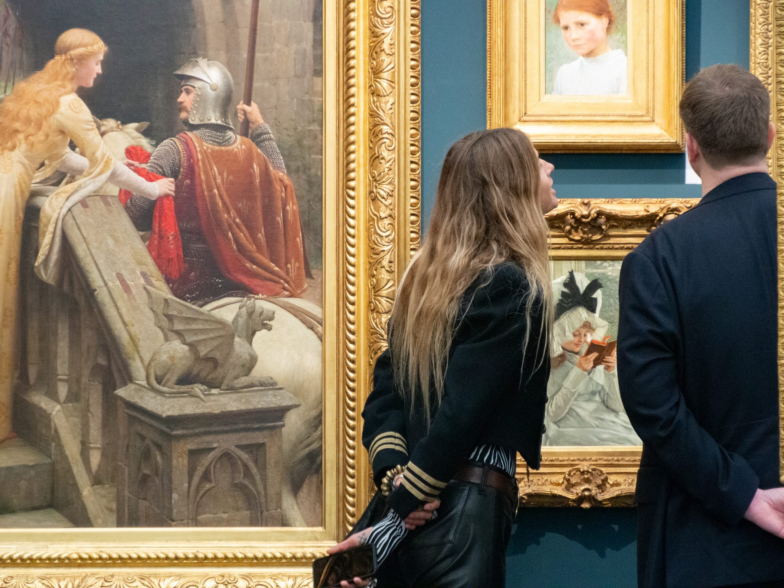 A young woman and man look at paintings in gold frames hung on a blue wall