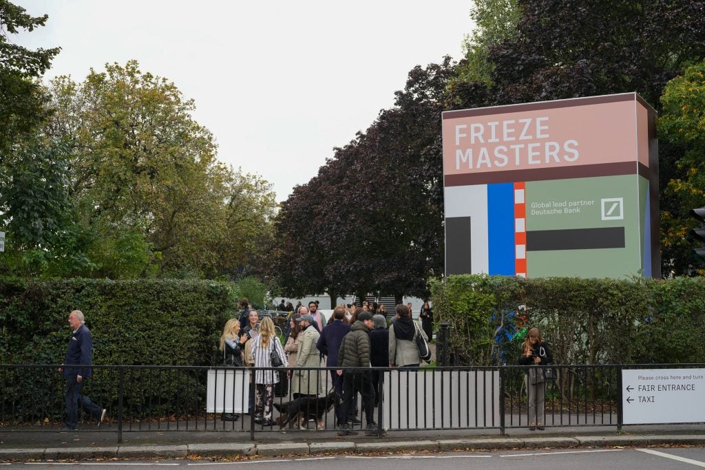 Visitors arriving at the entrance of Frieze Masters art fair in London, featuring the event’s large sign and surrounding greenery.