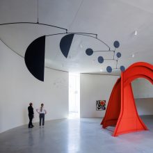 A photo of a spacious modern gallery with smooth white walls featuring a large black hanging mobile with circular elements and a towering red steel sculpture with curved arches, as two visitors stand observing the artworks.