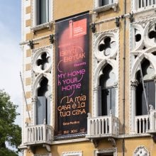 A large vertical banner reading “My Home Is Your Home” in English, Arabic, and Italian hangs from the façade of a historic Venetian building with ornate Gothic windows and small balconies. The banner advertises the Qatar Pavilion at the Venice Architecture Biennale, promoting free entry to the exhibition.
