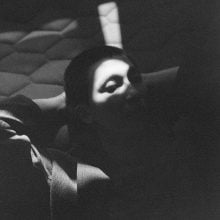 Moody black-and-white portrait of RM reclining, face partially obscured by dramatic shaft of light.