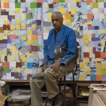 Artist seated in a studio wearing a paint-stained jacket, surrounded by colorful abstract square paintings, reflecting a creative and contemplative workspace.