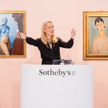 A woman in black blouse, blond hair, selling paintings at Sotheby's. She stands in the middle of two paintings hung on a coral wall.