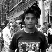 A black-and-white street photograph showing Jean-Michel Basquiat in the foreground wearing a Keith Haring T-shirt and a backwards cap. Behind him, people mill about on West Broadway in SoHo, New York, with storefronts, fire escapes, and a tilted "No Parking" sign visible.