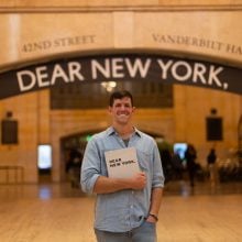 A photo of a smiling man in a light blue button-down shirt standing beneath an archway labeled “VANDERBILT HALL,” holding a book that reads “DEAR NEW YORK.”