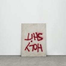 A canvas spray painted with words Holy Shit is displayed upside-down on the floor, leaning against the wall