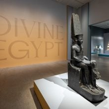 Installation view of the exhibition “Divine Egypt” at The Metropolitan Museum of Art, showing a large seated ancient Egyptian statue in the foreground and other sculptures displayed in softly lit galleries beyond.