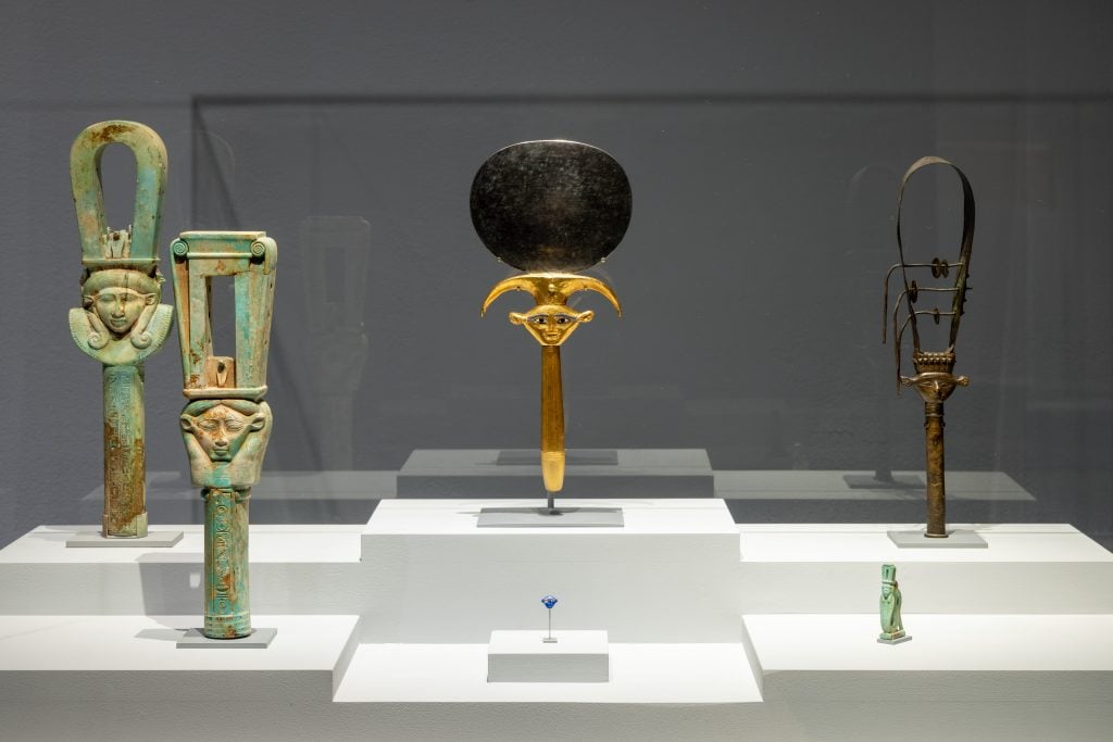 博物館展櫃裡陳列著五件古埃及文物，包括兩件飾有女神哈索爾面容的綠松石色叉鈴、一面飾有哈索爾圖案的金黑色禮儀鏡、一件繫有金屬絲帶的青銅叉鈴、一尊綠松石色小彩陶雕像，以及一件藍色小護身符。這些文物被精心佈置在灰色背景的白色階梯狀平台上，燈光照射得格外明亮。