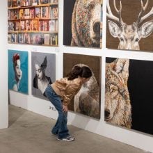 A child wearing a tan jacket and jeans bends forward to look closely at large animal-themed artworks, including a bear, deer, and wolf, displayed on a white gallery wall.