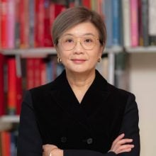An East Asian woman in glasses, black suit jacket, standing in front of a bookshelf.