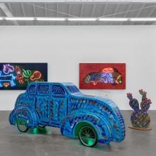 Exhibition scene with neon artworks of animals glowing on walls, a viewer observing a large blue sculptural car and a cactus-shaped sculpture in vivid hues.