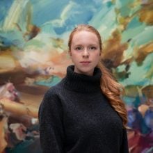 A photo of a young woman with fair skin and red hair, wearing a dark gray turtleneck sweater and standing before a large, colorful abstract floral painting.