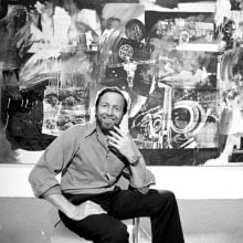 American artist Robert Rauschenberg (1925–2008) sits smiling in front of one of his large-scale mixed-media paintings during an exhibition of his work at the Museum of Modern Art, New York, March 1977. (Photo by Jack Mitchell/Getty Images)