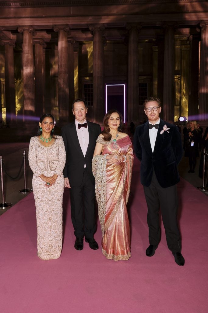 Four elegantly dressed guests posing on a pink carpet in front of a neoclassical building entrance illuminated at night.