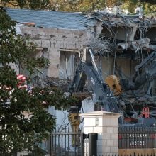 Excavators demolishing a partially collapsed building behind a metal fence, surrounded by trees and debris.