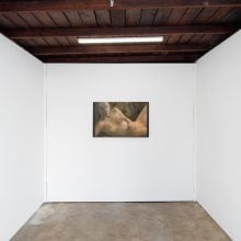 A minimalist gallery space with white walls and a wooden ceiling, displaying three paintings: a red monochrome on the left, a reclining nude at the center, and a colorful landscape scene on the right.