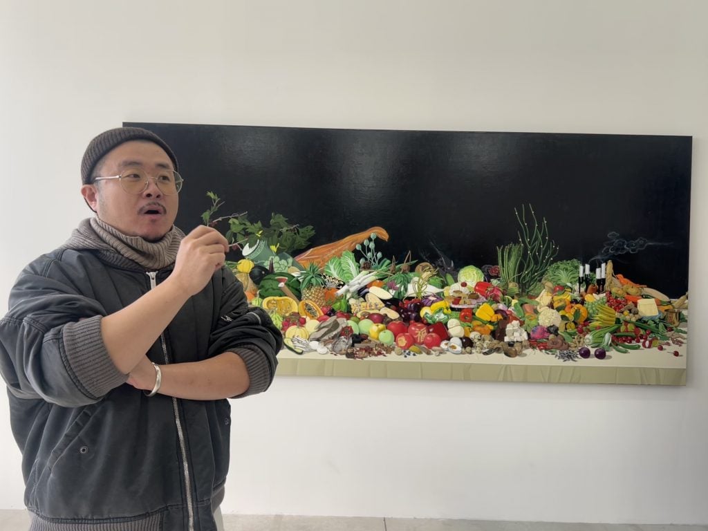 An asian man in a gray jacket and beanie stands in front of a still life of fruit