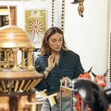 Kelly Wearstler stands in Pedro Friedeberg’s studio surrounded by eclectic artworks and objects. She wears a textured dark blouse and looks intently at a display of sculptural pieces, including red devil figures in glass-fronted boxes. Around her are ornate wooden constructions, chandeliers, and framed works with bold patterns and surreal imagery.