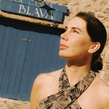 A woman, Spanish artist Berta De la Rosa, stands in warm sunlight before a blue wooden door marked “La Caseta Blava” in Mallorca. She gazes upward, her hair pulled back, wearing a patterned halter dress. The stone walls behind her evoke the rustic charm of Deià, where La Residencia, A Belmond Hotel, hosts the “Artists in Residencia” program.