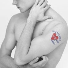 A black-and-white photo of a shirtless person with a tattoo on their arm reading “HEAD OVER HEELS AFTER MARX & ANGELS” inside a red heart.