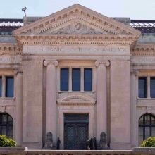 A photo of the facade of the Hispanic Society of America Museum and Library, an elegant Beaux-Arts building with tall Corinthian columns, arched windows, and a triangular pediment decorated with classical carvings. The stone exterior is inscribed with names of notable Spanish figures, including Cervantes and Lope de Vega. Two stone lion statues flank the entrance, adding to the building’s stately and monumental presence.