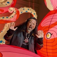 Takashi Murakami poses playfully with hands raised in front of a large, brightly colored installation featuring whimsical pink and red cartoon-like forms with bold swirling patterns and oversized eyes