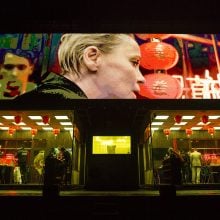 Theatrical set from ROHTKO with neon-lit restaurant below giant screen showing close-up faces in vivid colors.