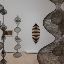 An installation view of Ruth Asawa’s hanging wire sculptures, their layered, bulbous forms suspended at varying heights, with a woven wall piece and framed drawings visible in the background.