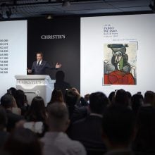 A man in suit auctioning a red and green Picasso cubism painting in a Christie's saleroom full of people.
