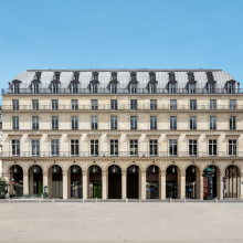 A photo of a grand neoclassical building in Paris with a symmetrical façade, tall arched ground-floor openings, and rows of evenly spaced windows topped by dormers with mansard roofs; the scene is taken from a wide plaza bordered by elegant historic architecture and a nearby café terrace.