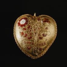 a golden heart-shaped pendant with a Tudor rose and pomegranates on it set against a black background