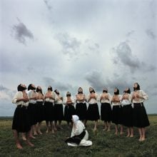 women standin a row holding their breasts, they are outside under grey skies