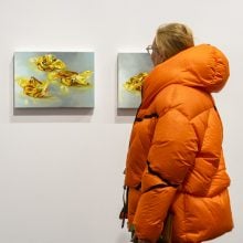 At Art Cologne 2024, a person wearing a bright orange puffer jacket observes three small paintings on a white gallery wall. Each painting depicts pieces of yellow and orange candy wrapped in translucent wrappers, arranged against soft blue-gray backgrounds.