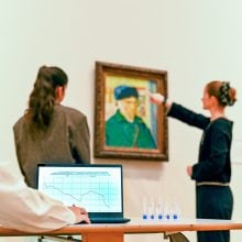 In the foreground, a computer screen, and in the background, two women stand in front of a Van Gogh self-portrait with bandaged ear
