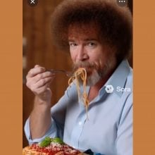 A man resembling Bob Ross, with curly hair and beard, eats spaghetti from a paint palette, wearing a blue shirt in a warmly lit room.