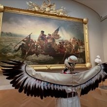A photo of an Indigenous woman seen from behind, wearing a white beaded robe with wide feathered wings outstretched, standing before the large painting “Washington Crossing the Delaware” in a museum gallery.