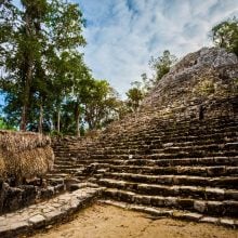 Ancient Maya pyramid at Cobá, Mexico, rises steeply from the jungle floor, its stone steps weathered by centuries. A small thatched-roof structure stands in the foreground, surrounded by carved stones and dense tropical vegetation.