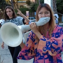 a photo shows protesters in the street. A woman at center holds a bullhorn that has been modified with a sculpture of a hand that covers her mouth