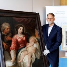 A photo of a middle-aged white man in a navy suit and white gloves standing with his hand resting on a large framed Baroque-style painting of two women and a child, with another framed artwork visible beside him in front of a Bavarian police banner.