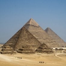 Three Giza pyramids rise from desert sands as camel riders traverse the foreground under clear sky.