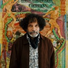 A Black man, artist Hew Locke, poses before his colorful artworks. He wears a dark jacket over a patterned shirt, has a salt-and-pepper beard and curly black hair.