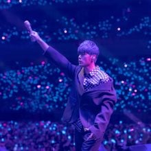Taiwanese singer Jay Chou holds a microphone out high with his right hand on a stage before a blue-tinged stadium filled with people.